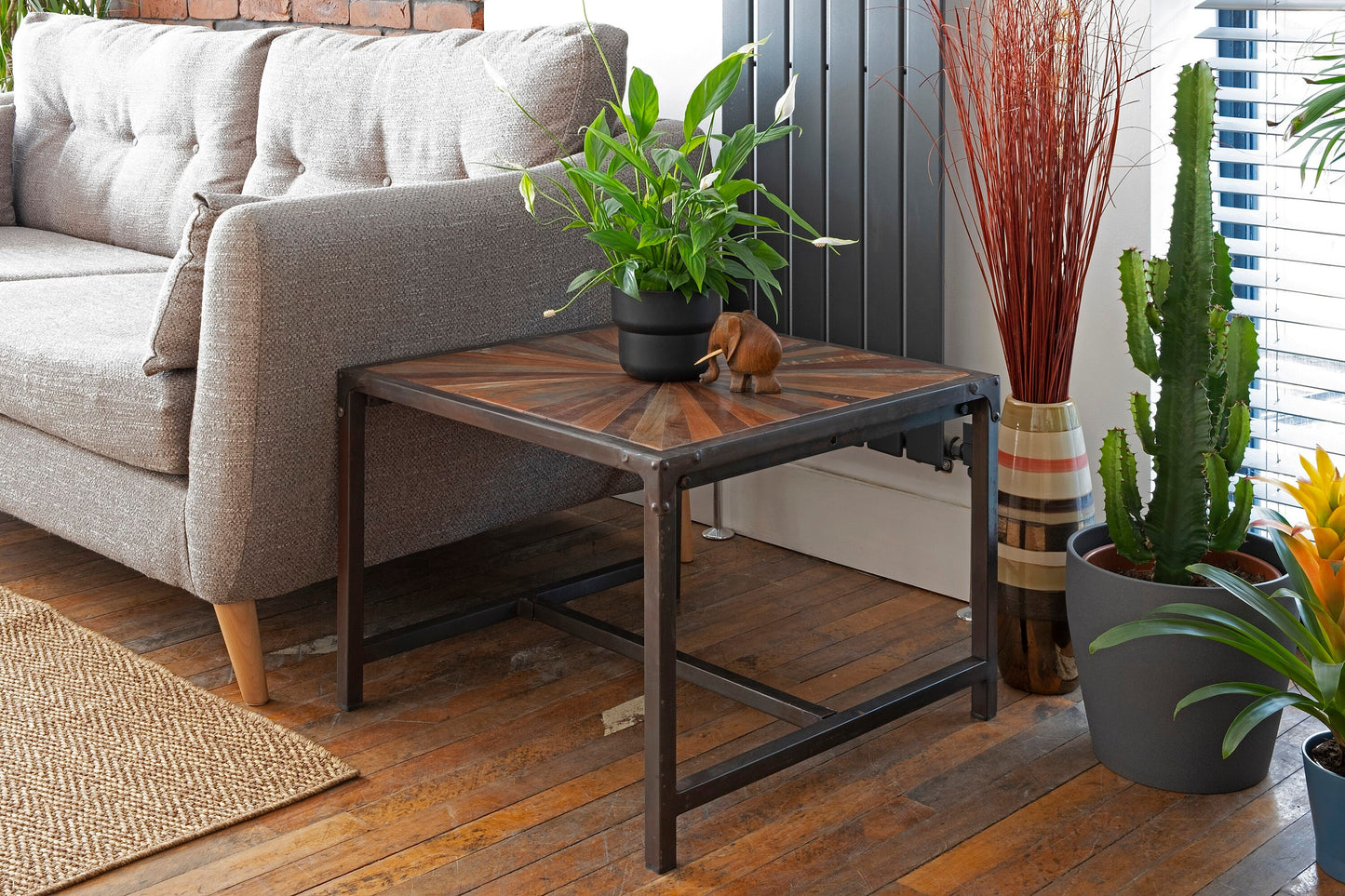 A square reclaimed teak table with a striking star-patterned, hand-painted distressed top sits beside a grey sofa, and has black iron legs, blending rustic and industrial style. A plant and wooden elephant sits on top, surrounded by indoor plants.