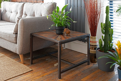 A square reclaimed teak table with a striking star-patterned, hand-painted distressed top sits beside a grey sofa, and has black iron legs, blending rustic and industrial style. A plant and wooden elephant sits on top, surrounded by indoor plants.