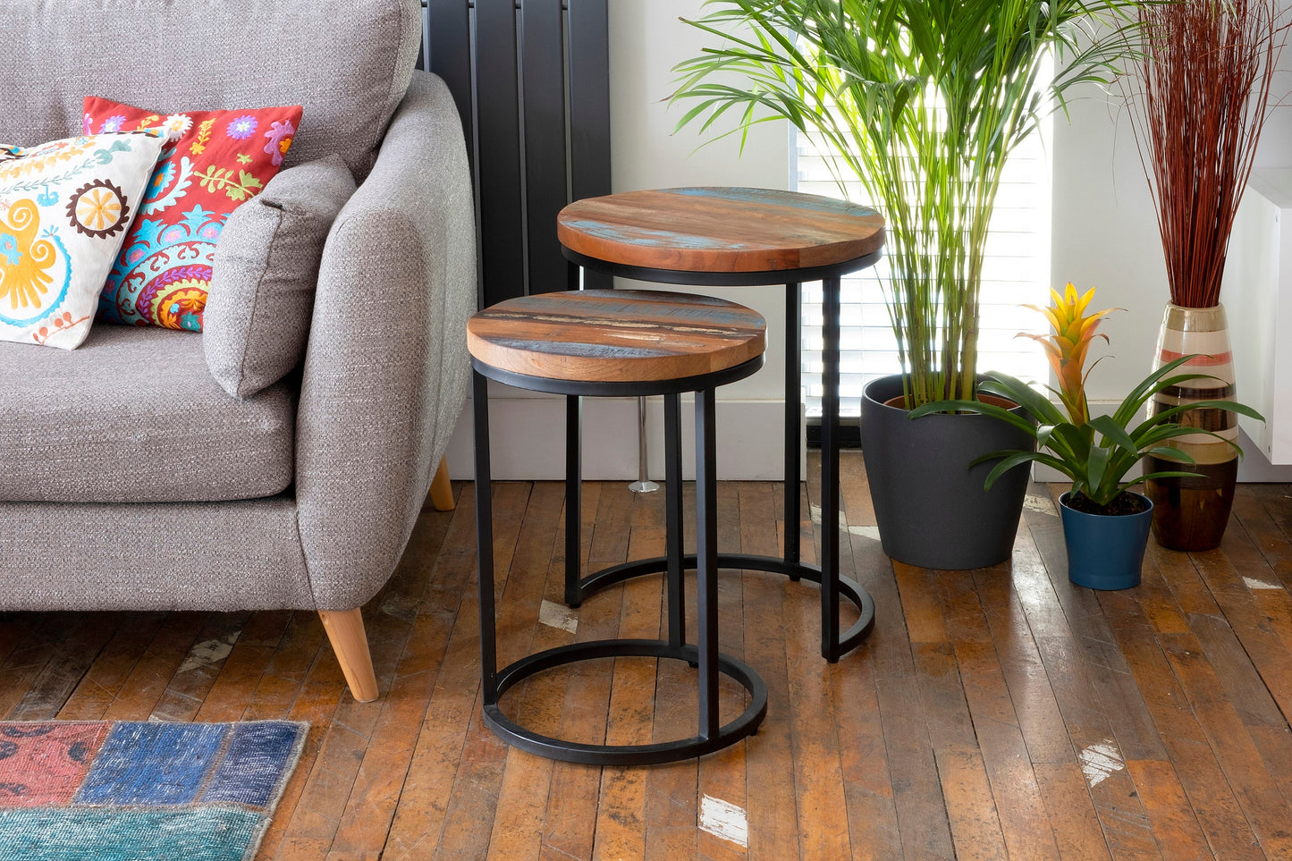 Two round, rustic wood and metal nesting tables with unique wooden tops, placed next to a gray sofa and colorful cushions. The tables are set on a wooden floor with potted plants beside them